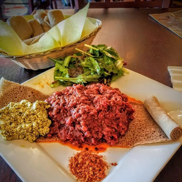 Ethiopian kitfo, gomen, and injera on a white plate, with a side salad and bread basket.