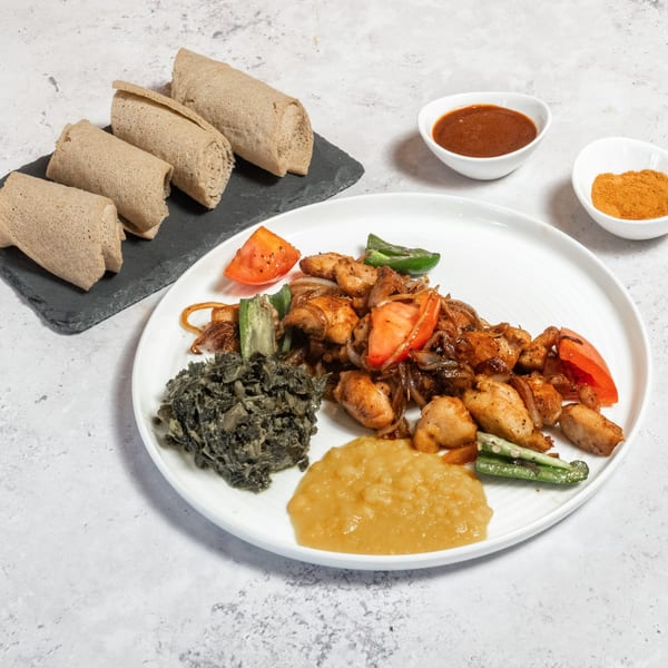 Ethiopian chicken tibs with injera, collard greens, and sauces on a white plate.