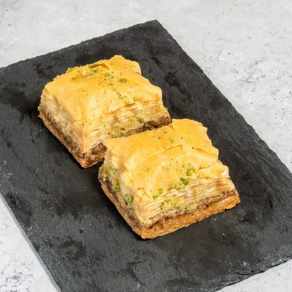 Two pieces of golden baklava with pistachio sprinkles on a dark slate board.