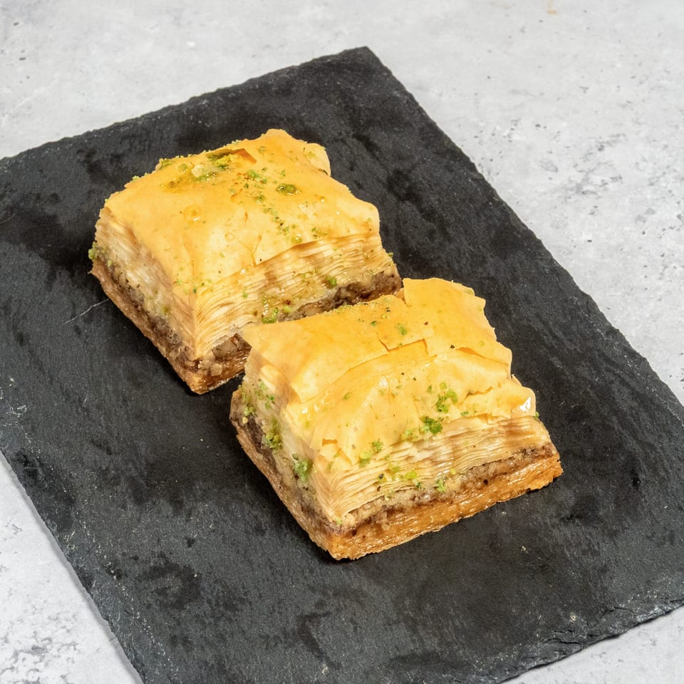 Two pieces of golden baklava with pistachio sprinkles on a dark slate board.