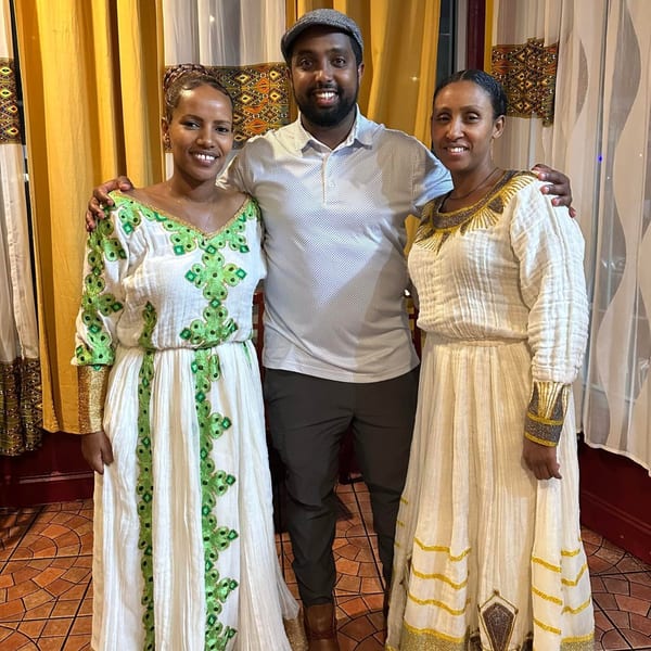 Three smiling people, two women in traditional Ethiopian dresses and a man in a polo shirt.